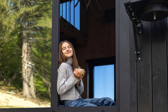 Young Woman Enjoying Coffee by Cabin Window. Young woman relaxing with a cup of coffee by a large window in a modern forest cabin.
