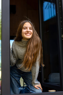 Smiling Young Woman by Cabin Window. Smiling young woman leaning through a modern cabin window in a cozy natural setting.