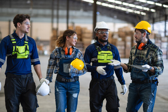 Stacks flank factory workers discussing teamwork as four colleagues walk through a team meeting while carrying a tablet.