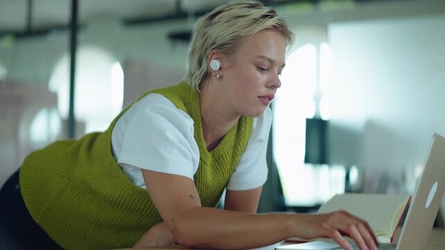 Woman talks and types on a laptop during an online meeting at a desk, using earbuds and a notebook for remote work, freelance communication, and productivity.
