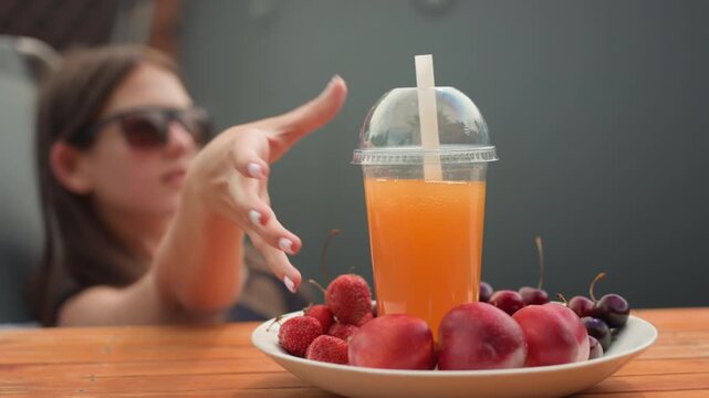 Young woman reclines on chair reaching for and holding orange drink with straw. plate of fresh fruit including peaches cherries and strawberries sits nearby on wooden table in soft natural