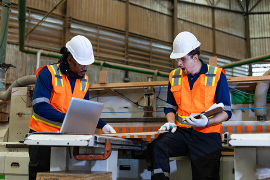 Worker fatigue: a tired factory worker yawns with a clipboard as his colleague reviews a laptop at a panel saw.