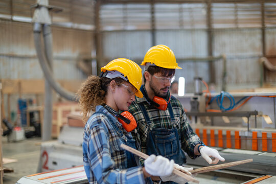 Female carpenter: woman in a carpentry workshop holds wood while her male colleague points at the piece beside a panel saw.