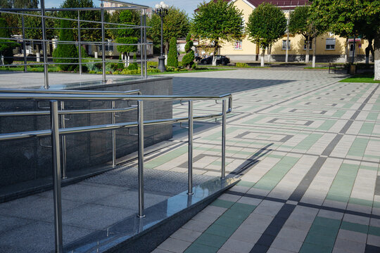 Outdoor concrete ramp with stainless steel shiny handrails leading to public square with paving tiles on sunny summer day