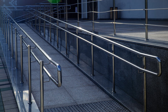 Accessible building entrance ramp made of stone tile with shiny stainless steel handrails for wheelchair users on sunny day