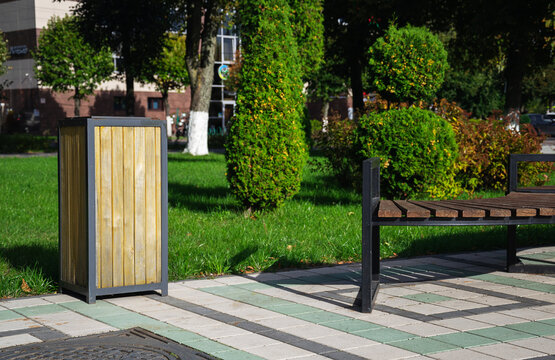 Wooden waste bin stands near park bench on tiled pavement of public square surrounded by green lawns and trees on sunny day