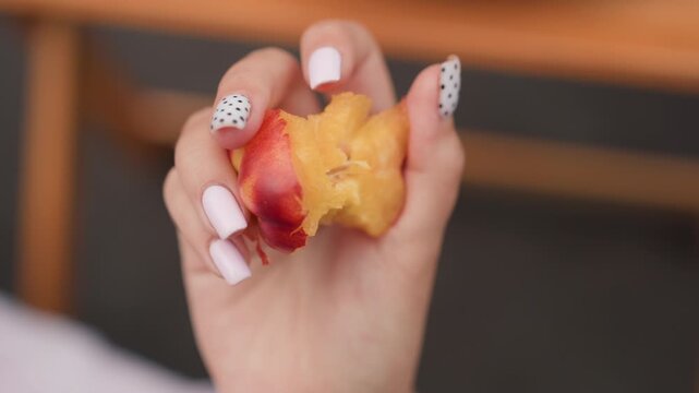 Hand with white and polka dot nail polish holds partially eaten peach displaying fruit texture. background remains softly blurred throughout emphasizing close up view of food and nails