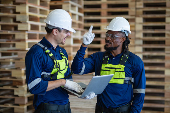 Discussing wooden pallet logistics in a warehouse, two men confer as one points upward and the other holds a laptop.