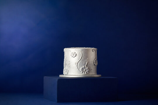 A silver, decorated cake is displayed on a dark pedestal against a deep blue background.
