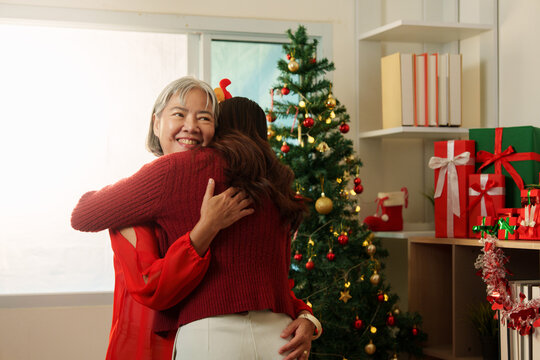 Senior Asian mother embracing adult daughter by Christmas tree for seasonal family reunion