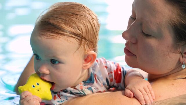 Cute baby biting yellow star toy while resting on mothers shoulder in indoor pool