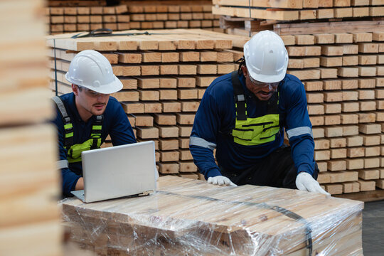 Two warehouse colleagues review pallet logistics using a laptop resting on wrapped lumber atop a loaded wooden pallet.