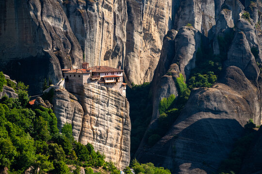 Aerial view of the Meteora monastery perched on a high rock formation, surrounded by dramatic cliffs, lush green trees, and deep shadows under a clear sky in Trikala, Greece.
