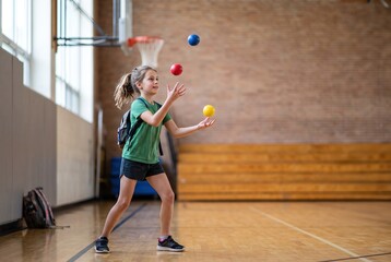 Fototapeta premium A girl with a backpack practices juggling colored balls in a gym. She smiles as she focuses on her activity. Other children are nearby, engaging in different leisure activities