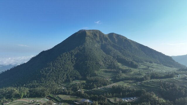 Aerial view of Mount Andong with lush green slopes, terraced fields, and clear blue sky in Kecamatan Ngablak, Jawa Tengah, Indonesia.