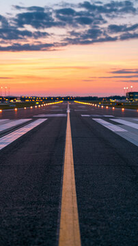 Empty runway at an airport in the evening with amber light grazing the asphalt centerline markings receding beneath a rose colored sunset sky distant edge lights beginning