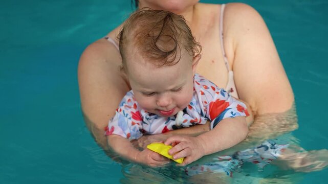 Cute infant holding yellow toy while resting in mothers arms in pool