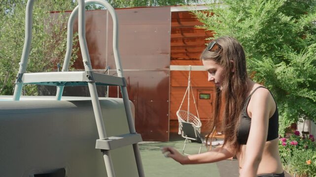 Young woman in black sports bra and skirt climbs out of above ground pool adjusting her position on ladder. scene is set outdoors with green trees bright sunlight garden flowers wooden