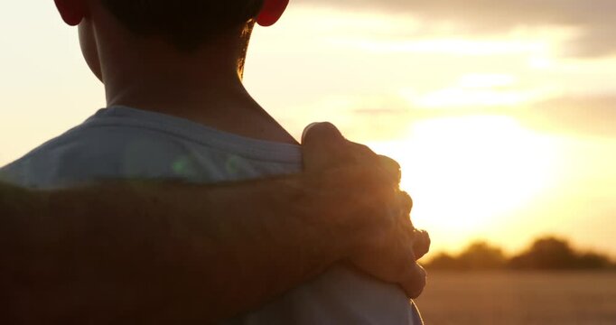 Father rests hand on son's shoulder as they watch sunset over field. Child and parent share quiet outdoor moment in golden light. Family support and love create hope for lifestyle storytelling.