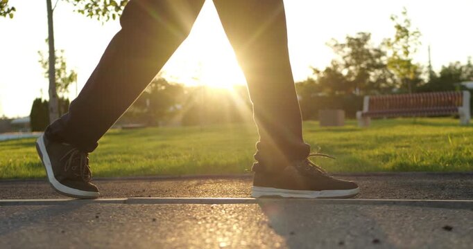 Person walks on park path in black sneakers at golden hour. Close-up footsteps show outdoor exercise and active lifestyle. Sunset light creates peaceful wellness background for fitness concepts.