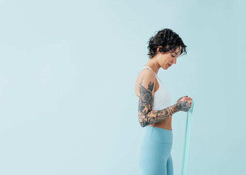 Side view of a slim woman in sportswear practicing with a resistance band over a blue backdrop