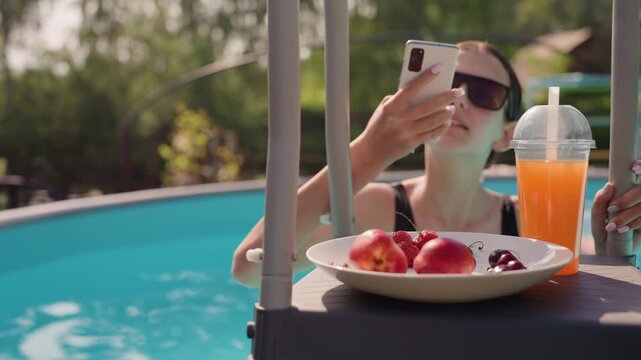 Woman relaxes by pool taking selfies with her smartphone while enjoying fresh fruit and orange juice. Woman wears sunglasses in bright outdoor lighting near clear blue water creating casual