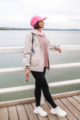 Full length side view of a happy millennial solo traveler in pink cap looking at the sea, smiling female tourist in windbreaker jacket and leggings on a pier © MarijaBazarova