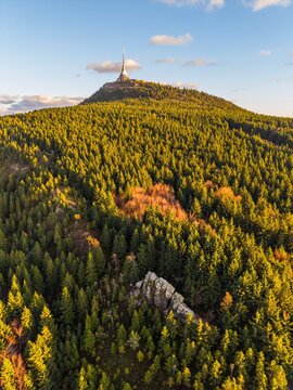 Aerial view of Jested Mountain and its tower above dense coniferous forest with a rock formation in the foreground during golden hour in Svetla pod Jestedem, Liberec Region, Czechia.