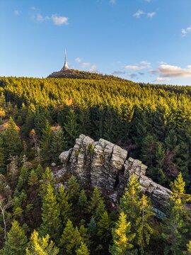 Aerial view of the Stone Gate rock formation surrounded by dense forest with the Jested Tower on the mountain peak under a clear blue sky in Svetla pod Jestedem, Liberec Region, Czechia.