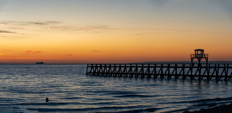 View of a wooden pier in front of the sea, a colorful sunrise, a cruise ship and port of Le Havre far behind