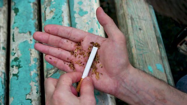 Man rolling a hand-rolled cigarette with loose tobacco in his palm against a weathered wooden bench background
