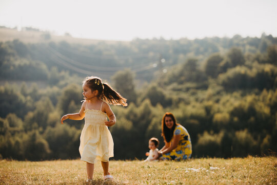 Little Girl Running in Sunny Meadow While Mother Watches With Baby During Summer Family Play
