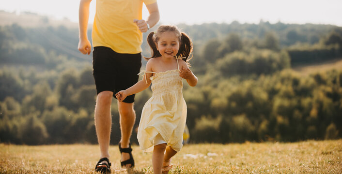 Father Running With Young Daughter Across Sunlit Field On Summer Afternoon