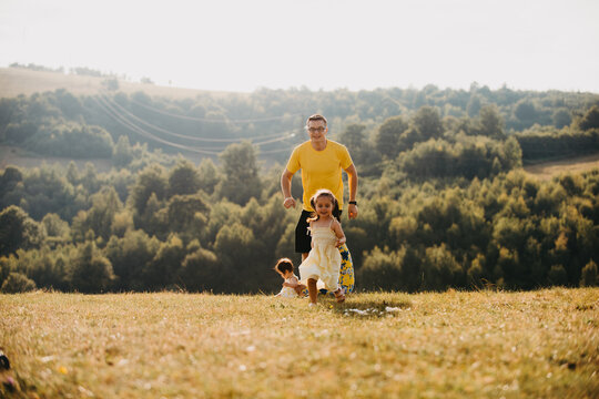 Father And Daughters Running Together In Sunny Summer Meadow With Forested Hills