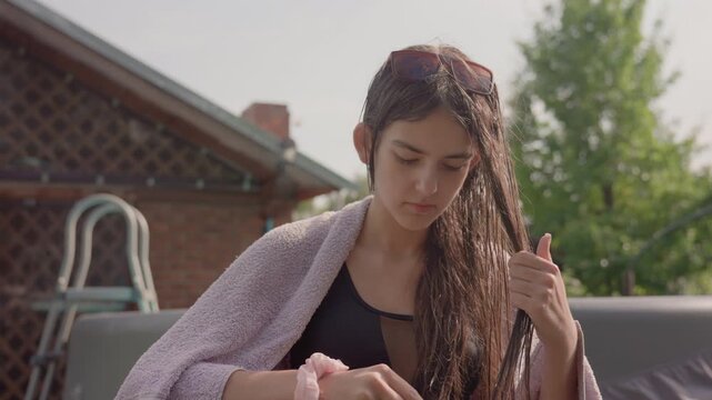 Young woman with wet hair combs her long brown locks while wearing black swimsuit and light purple towel outdoors near brick house and greenery. Woman holds comb in one hand and lets loose