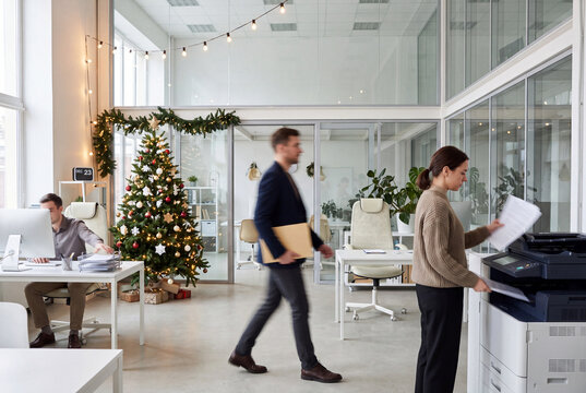 Employees continue working in a modern office decorated for Christmas with a lit tree and garlands; the active workplace conveys pre-holiday bustle as daily business continues in a festive setting.