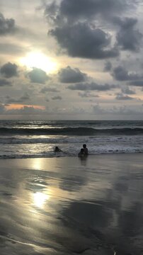 Two people standing on a beach at sunset. The ocean waves gently lap at the shore. The sky is filled with clouds and a warm glow from the setting sun.