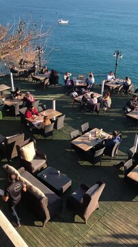 Outdoor dining area with tables and chairs overlooking the ocean. People are enjoying their meals in a relaxed atmosphere. A boat is visible in the distance.