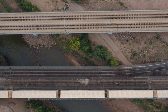 Aerial view of two parallel railway bridges crossing a river with a freight train passing over one of them, surrounded by trees and a path in Denver, Colorado, United States.