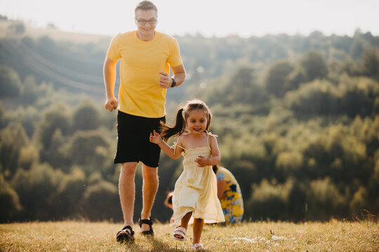 Father Chasing Daughter Through Sunlit Meadow During Summer Family Outdoor Playtime And Joyful Moments