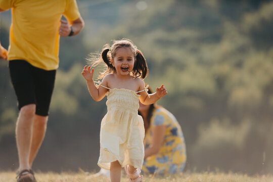 Happy Little Girl Running Toward Camera During Family Picnic In Sunlit Meadow With Parents