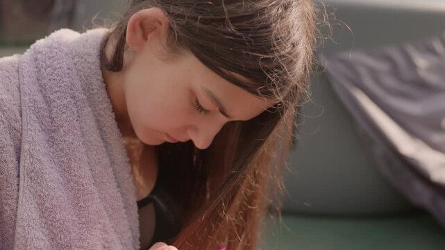 Young girl combs her wet hair with pink comb while wearing purple towel and light pink scrunchie focusing intently on grooming process in outdoor setting