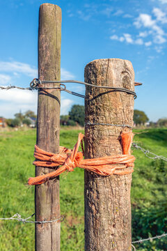 Close-up of rough wooden posts connected by iron wire and orange rope. The posts stand in the foreground of a rural area.