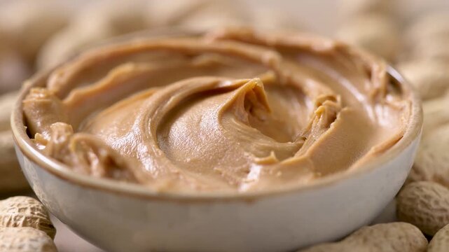 creamy peanut butter with a smooth decorative swirl on the surface, rotating on a rustic kitchen tabletop surrounded by scattered peanuts