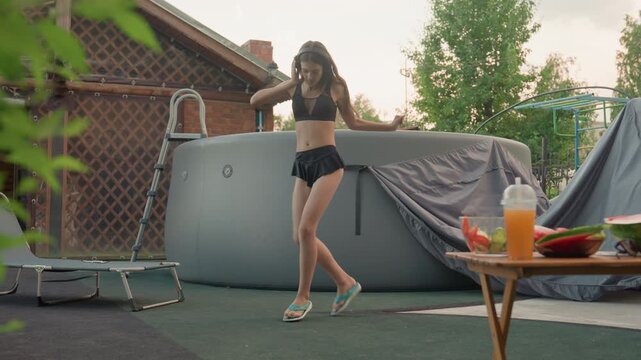 Young woman in black sports bra and skirt dances energetically near outdoor hot tub with flip flops on. table holds fruit slices and juice while background features greenery lattice fencing