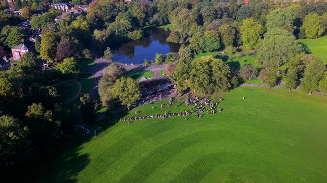 Aerial video looking over the historic pavilion, boating lake, open playing greens, and morning runners in Alexandra Park near Princes Road in Manchester. 