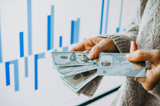 Hands holding US dollars currency and a bank card in front of the stock exchange screen showing trading graphics.