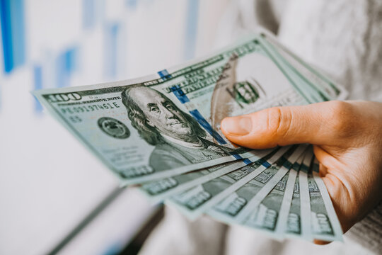 Hands holding US dollars currency in front of the stock exchange screen showing trading graphics.