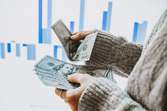 Hands holding US dollars currency in front of the stock exchange screen showing trading graphics.