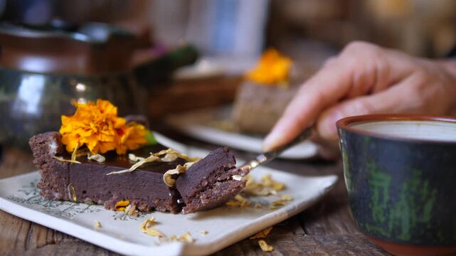 Fork serves dense chocolate cake on white plate next to cup, with marigold garnish and scattered crumbs on wooden table. Gentle hand movement suggests relaxed cafe dessert tasting and enjoyment.